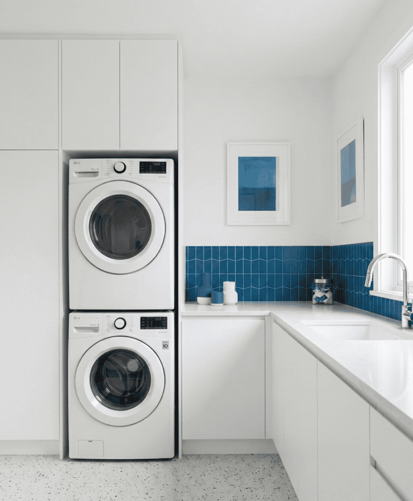 Crisp white minimalist laundry room featuring seamless handleless cabinetry, stacked washer and dryer integration, and vibrant blue tile accents. Custom space-saving utility millwork in White Rock, BC.